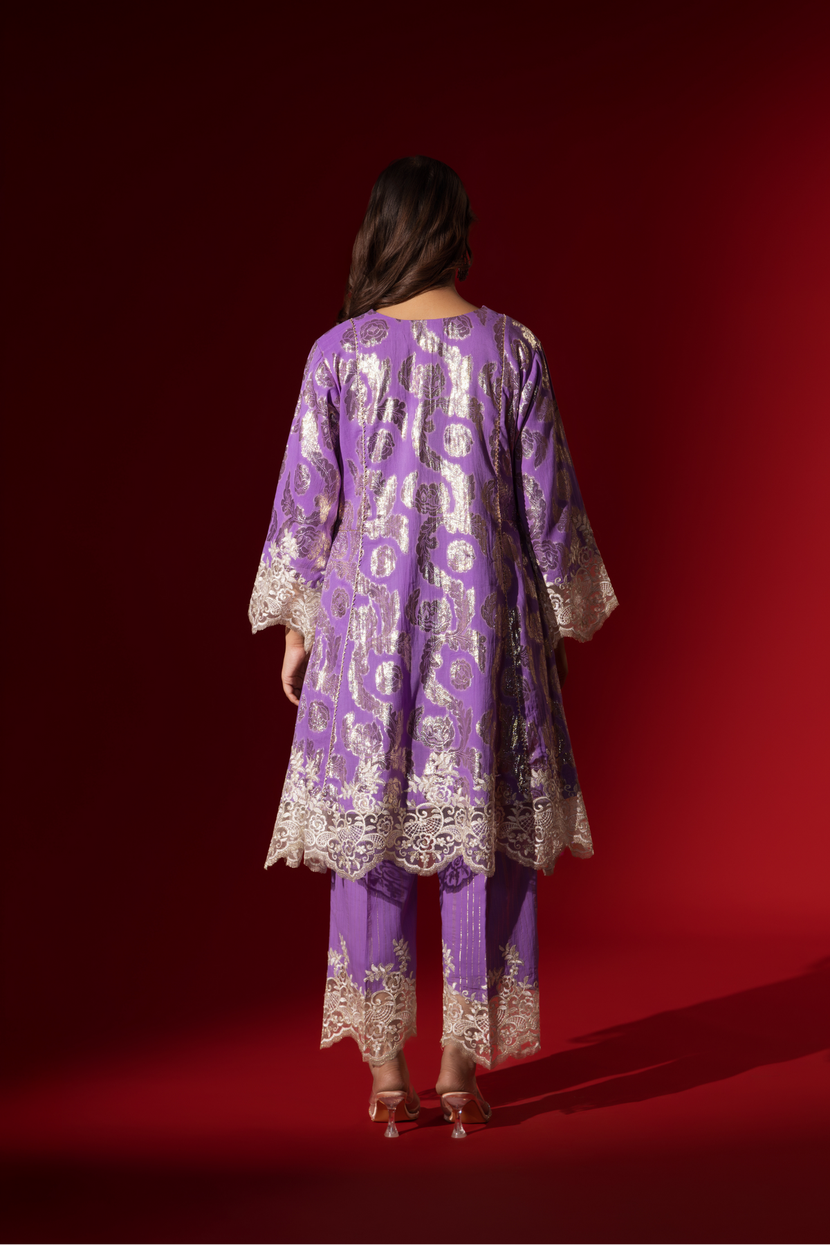 Back view of a woman poses in a purple ethnic shimmer kurta set outfit with intricate white embroidery against a vibrant red backdrop.