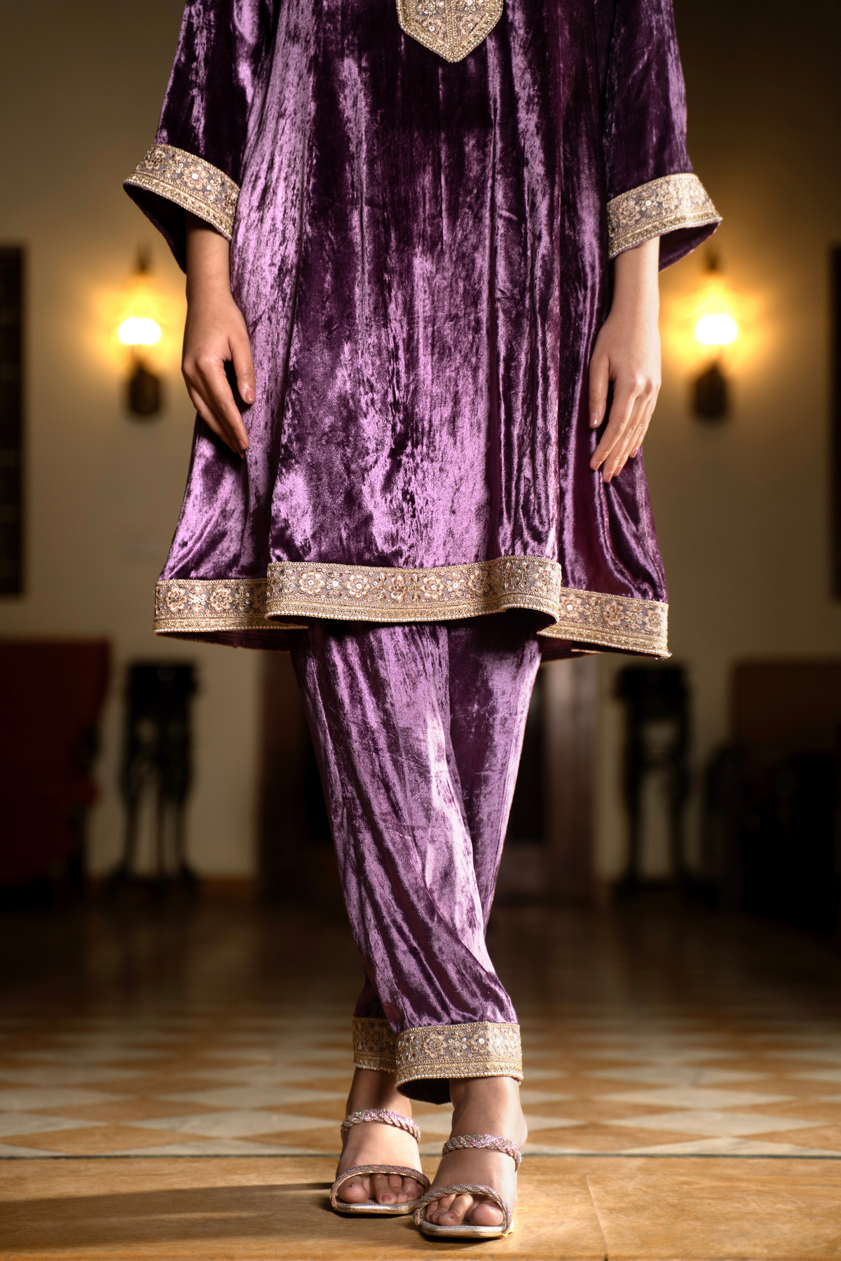 A woman stands elegantly in a purple velvet traditional outfit with embellished cuffs, showcasing her stylish shoes in a softly lit space.