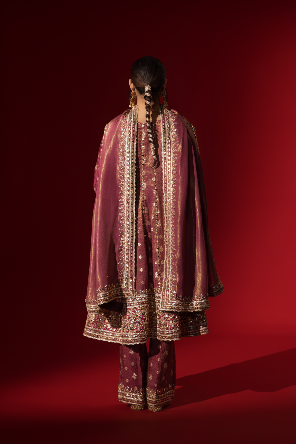 Back view of a woman stands elegantly in a rich maroon embroidered brocade kurta set adorned with intricate gold embroidery, against a deep red background.