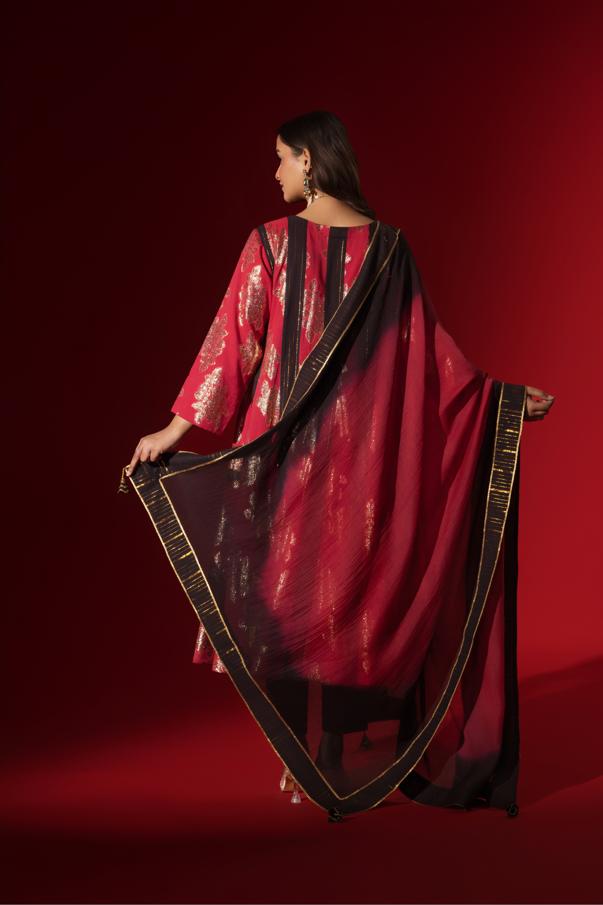 Back view of a woman poses in a vibrant red and black traditional outfit, adorned with intricate patterns and complemented by elegant jewelry.