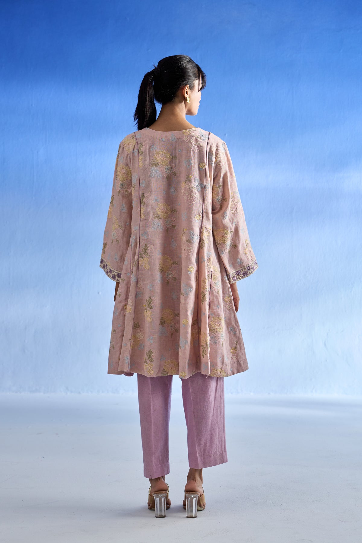 Back side of a woman shown from the back wearing a light pink linen kurta set with a subtle yellow floral pattern and lavender pants, standing against a blue gradient studio background.