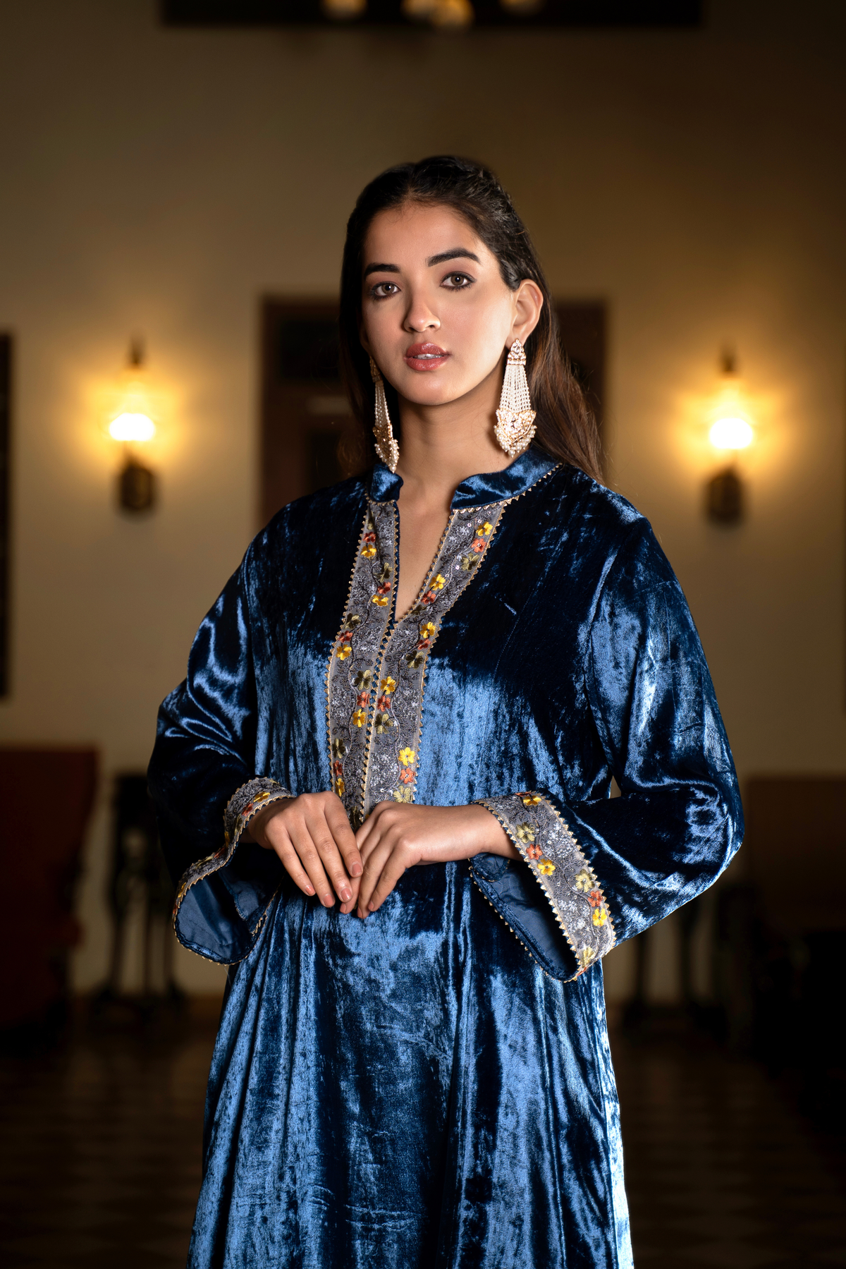 Closeup view of a woman stands in a traditional setting, wearing a vibrant blue velvet co-ord set outfit with decorative accents, surrounded by elegant furnishings.