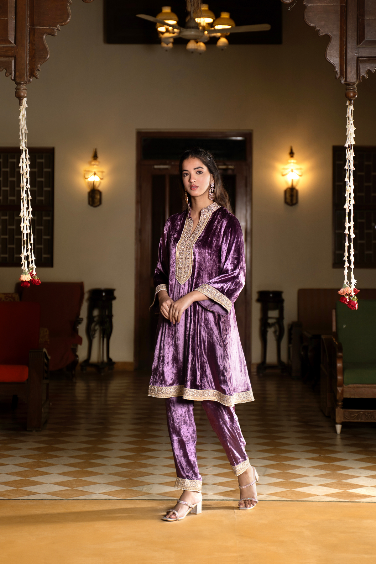 Side view of a woman stands confidently in a traditional purple velvet co- ord set outfit with intricate embroidery, set against a classic interior with warm lighting.