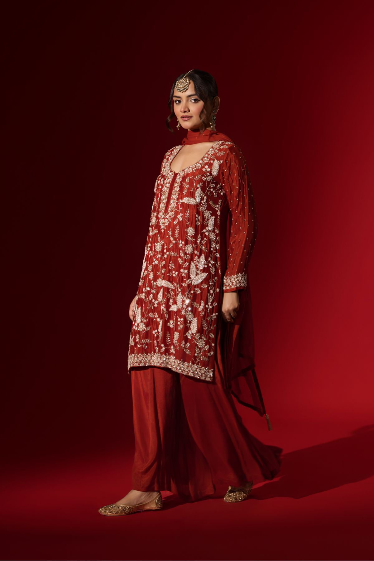 Side view of a woman poses in an elegant red embroidered kurta set adorned with intricate white embroidery against a rich red background.