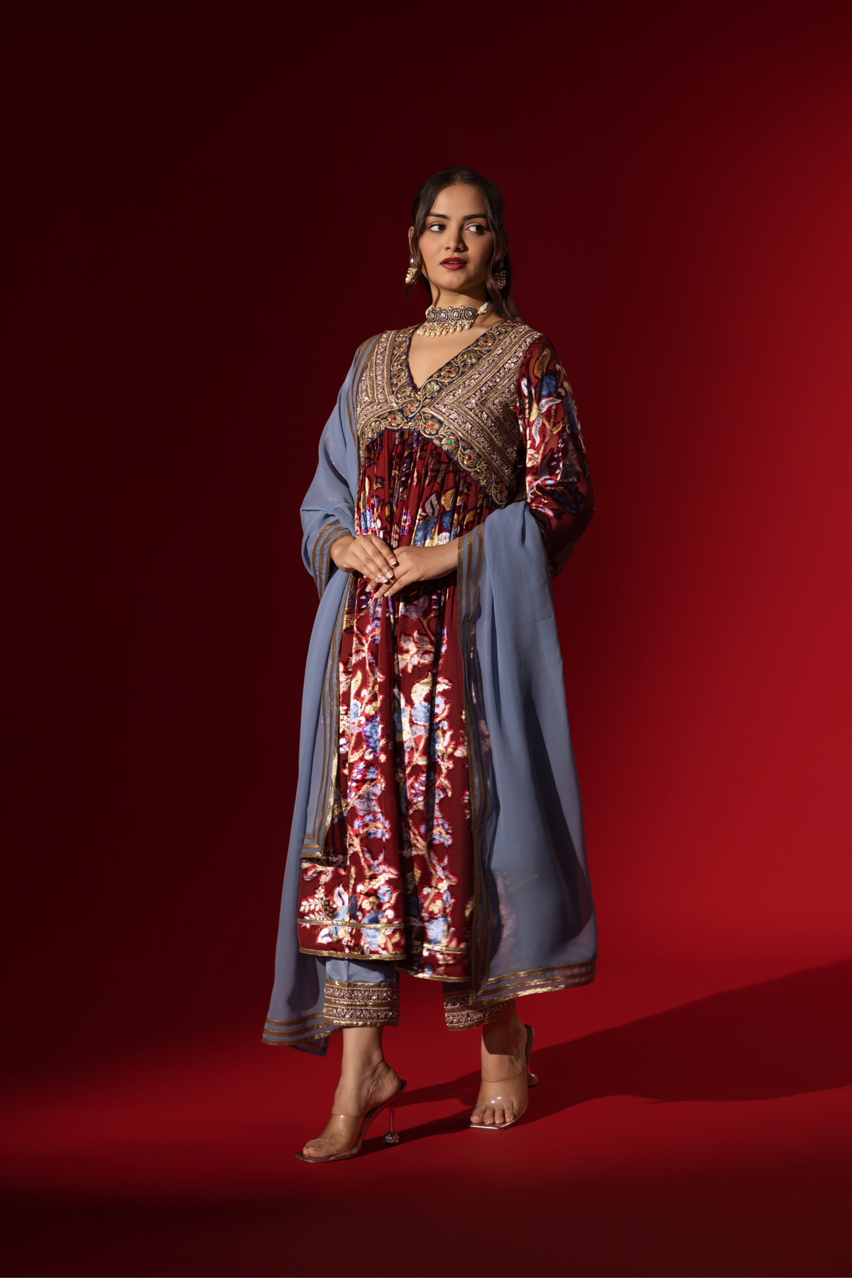 Side tilt view of a woman in traditional attire features a richly embellished maroon and floral dress, paired with a blue shawl, against a deep red background.