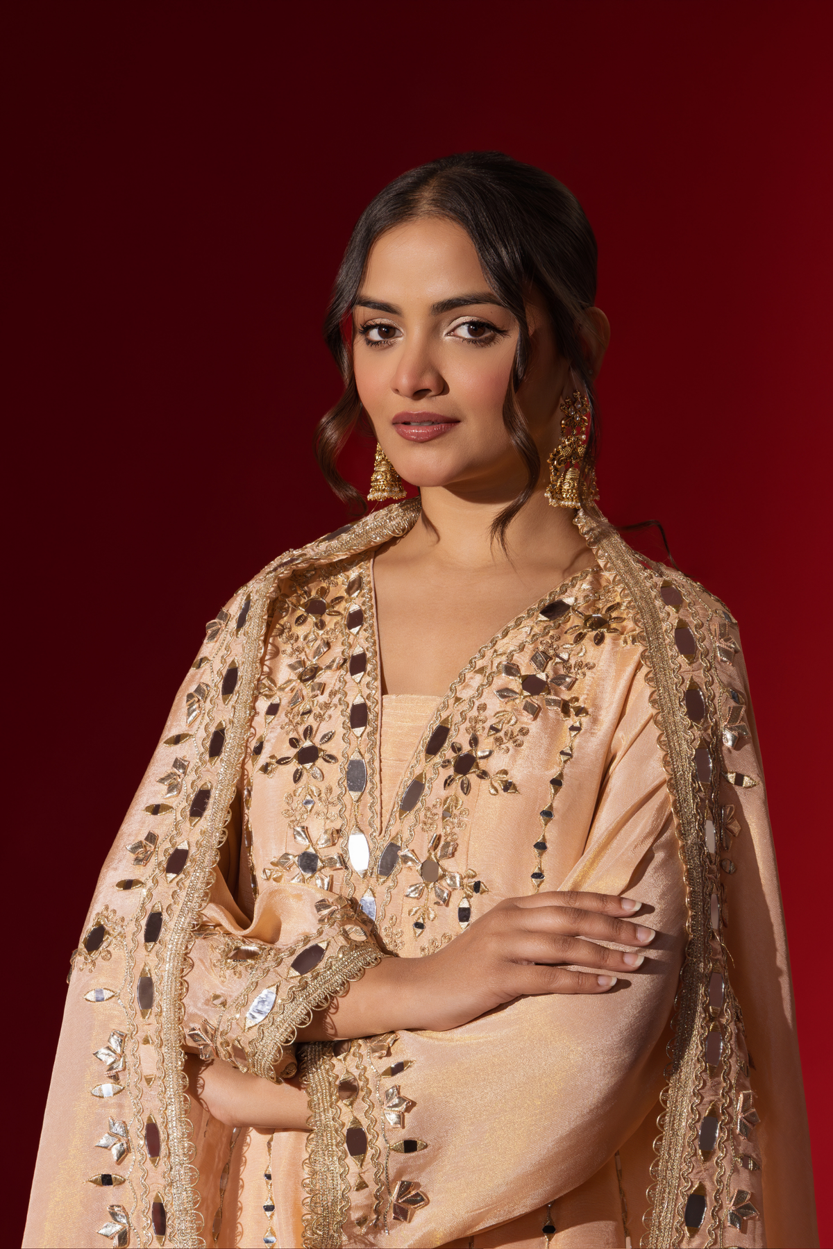Closeup view of a woman stands elegantly in a beige embroidered kurta set with intricate patterns, complemented by a matching dupatta against a bold red background.