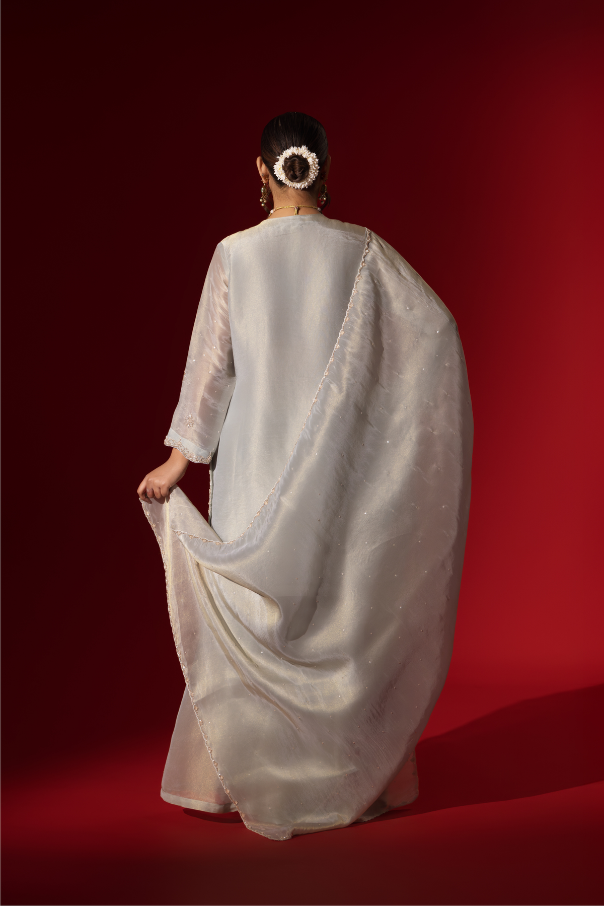 Back view of a woman stands elegantly in a shimmering silver grey embroidered gharara set, featuring intricate embroidery, paired with matching wide-legged trousers and a flowing shawl.
