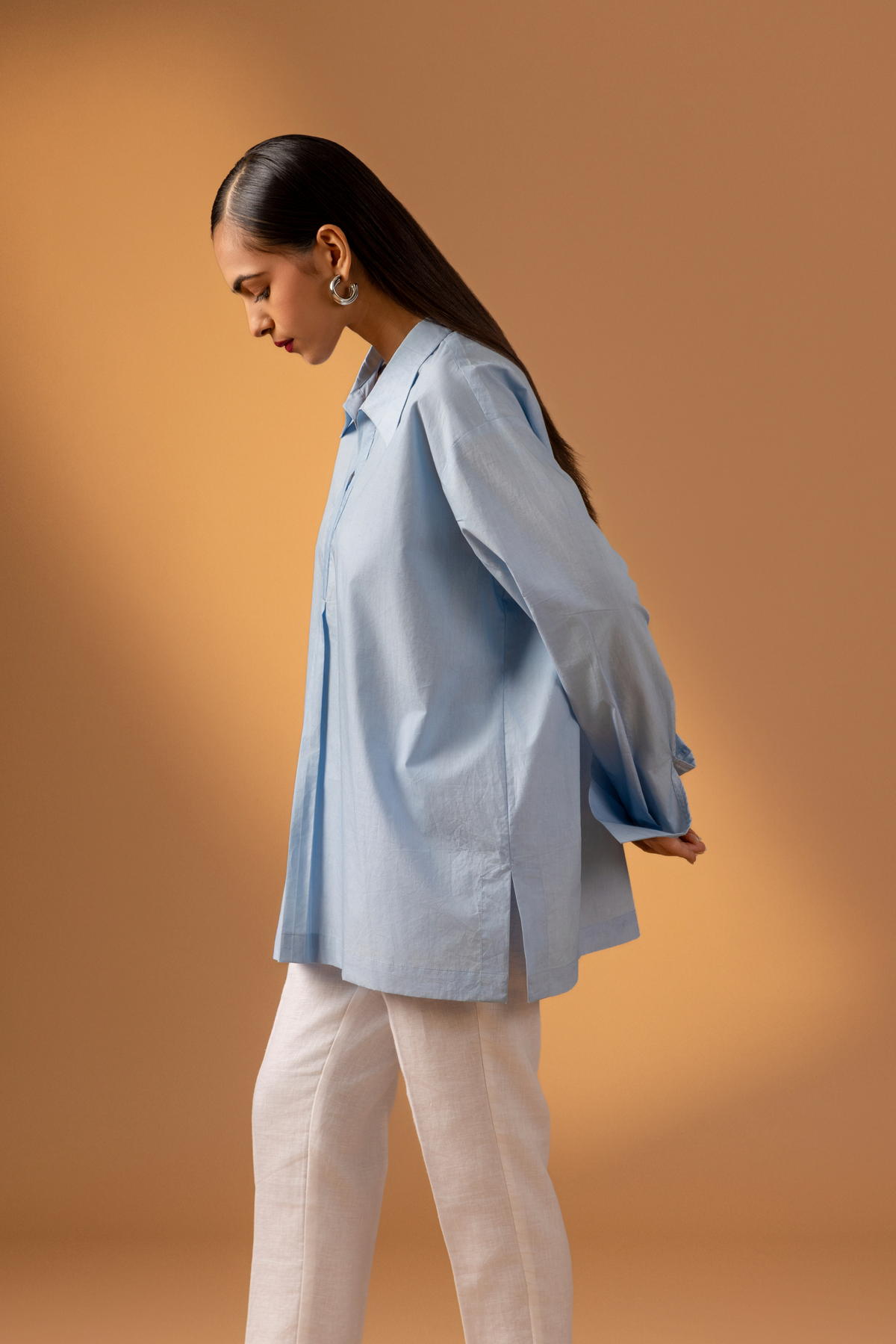 Side view of a women stands confidently wearing a light blue oversized shirt, white pants, and stylish sandals, against a warm beige background.