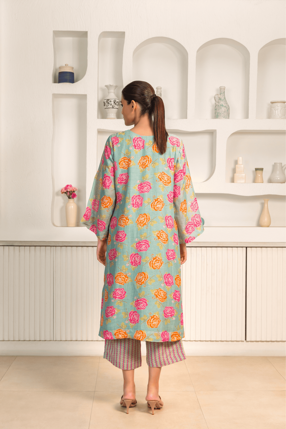 Back view of woman is wearing a light blue kurta with vibrant pink and orange floral prints, paired with patterned pants and posing against a white minimalist backdrop featuring built-in shelves with vases and decorative items.
