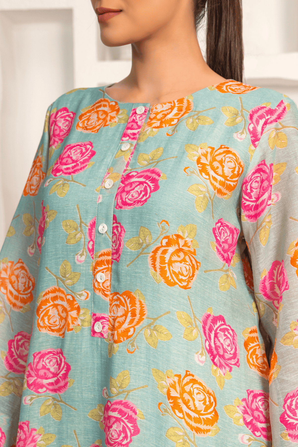 Closeup view of woman is wearing a light blue kurta with vibrant pink and orange floral prints, paired with patterned pants and posing against a white minimalist backdrop featuring built-in shelves with vases and decorative items.