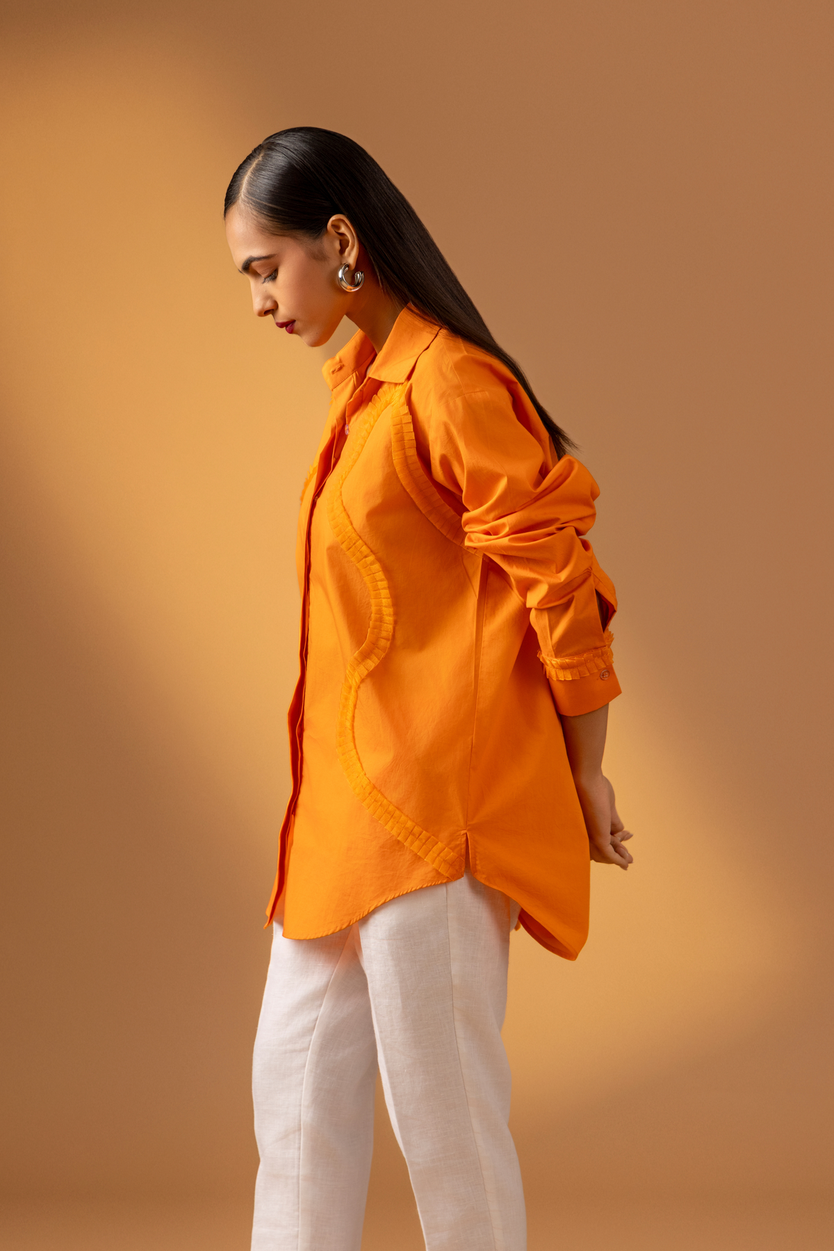 Side view of a woman stands confidently in an orange shirt paired with white trousers, and sandals, against a warm, neutral background.