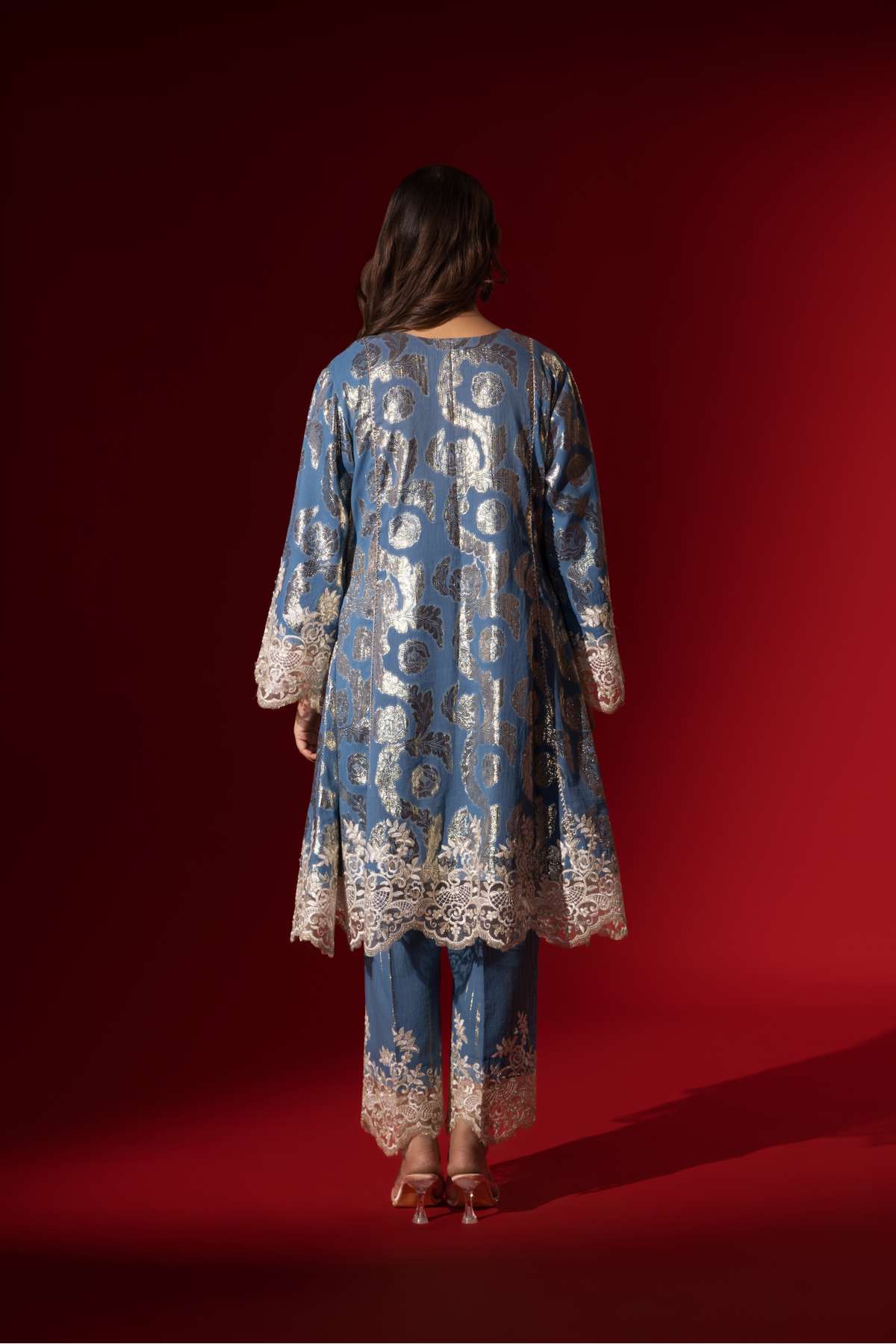 Back view of a woman poses in a blue ethnic shimmer kurta set outfit with intricate white embroidery against a vibrant red backdrop.