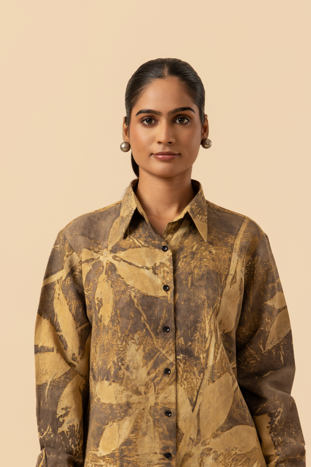 Closeup view of a model wearing a patterned brown and yellow shirt with beige wide-leg trousers and sandals, standing against a soft beige background.
