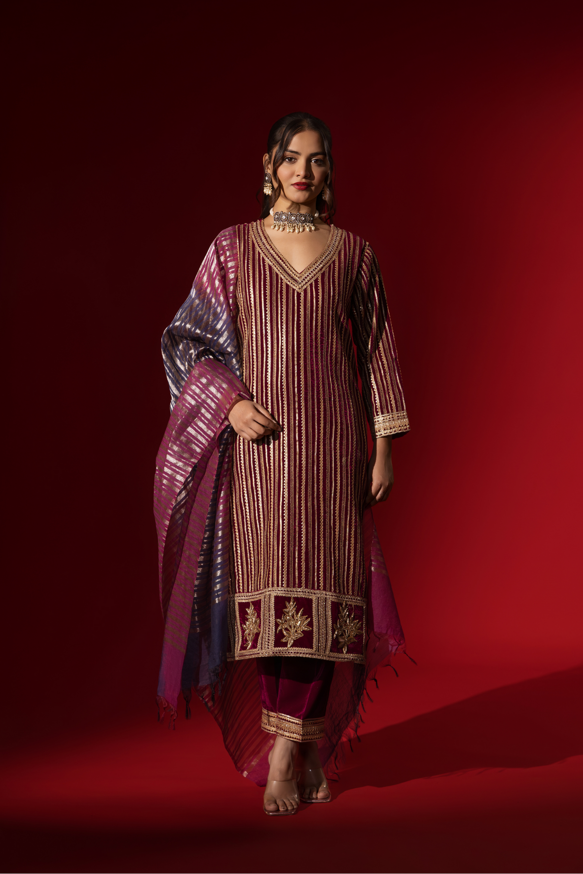 A woman showcases an ethnic wine velvet gota kurta set with golden stripes, paired with a shimmering shawl, against a rich red backdrop.