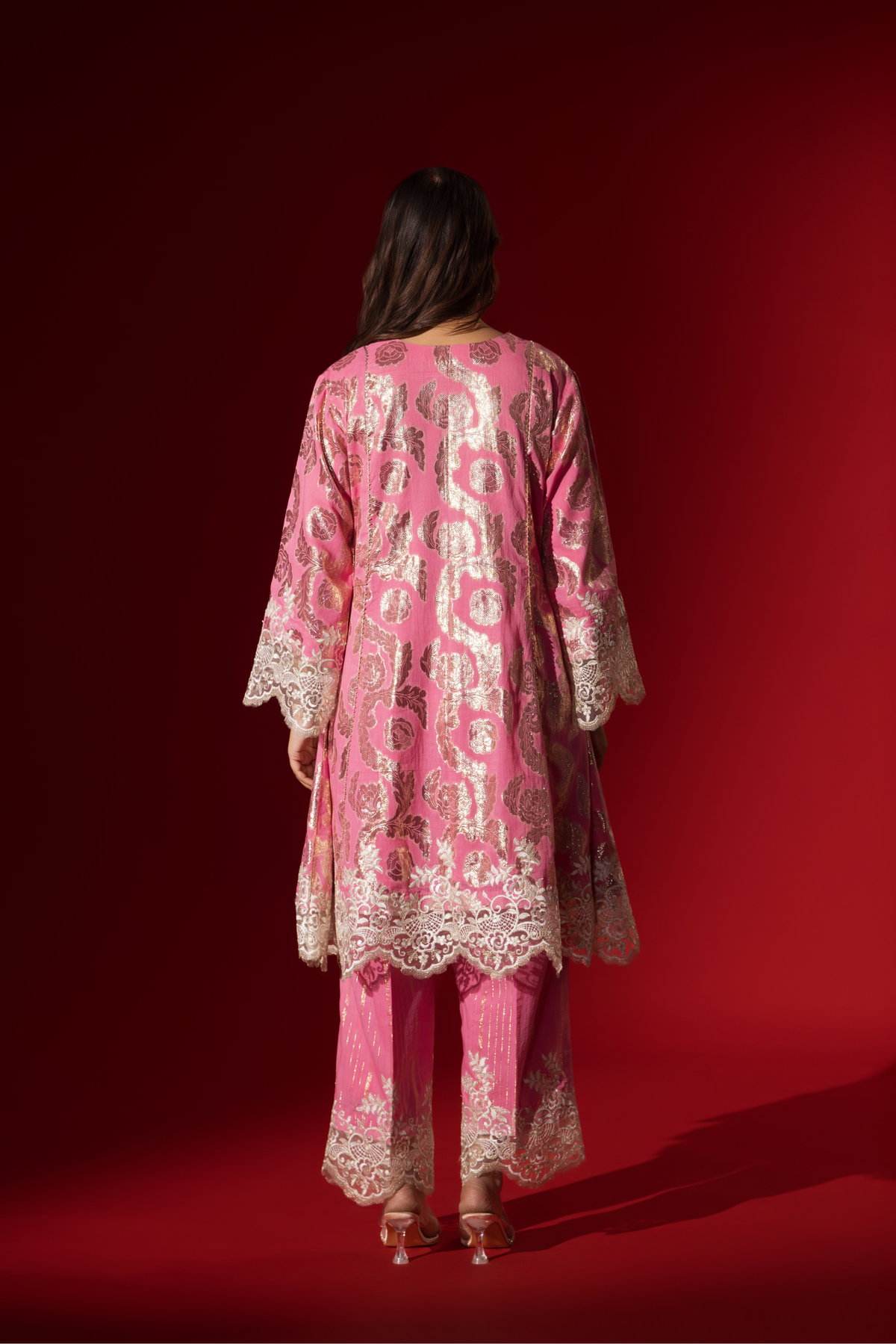 Back view of a woman poses in a pink ethnic shimmer kurta set outfit with intricate white embroidery against a vibrant red backdrop.