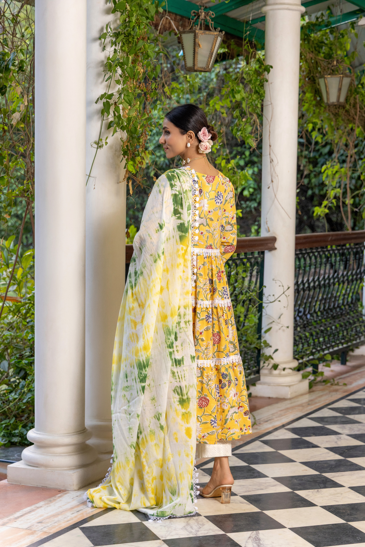 Back view of a woman stands gracefully in a vibrant yellow floral cotton kurta set, paired with a light green and white dupatta against a lush, green backdrop.