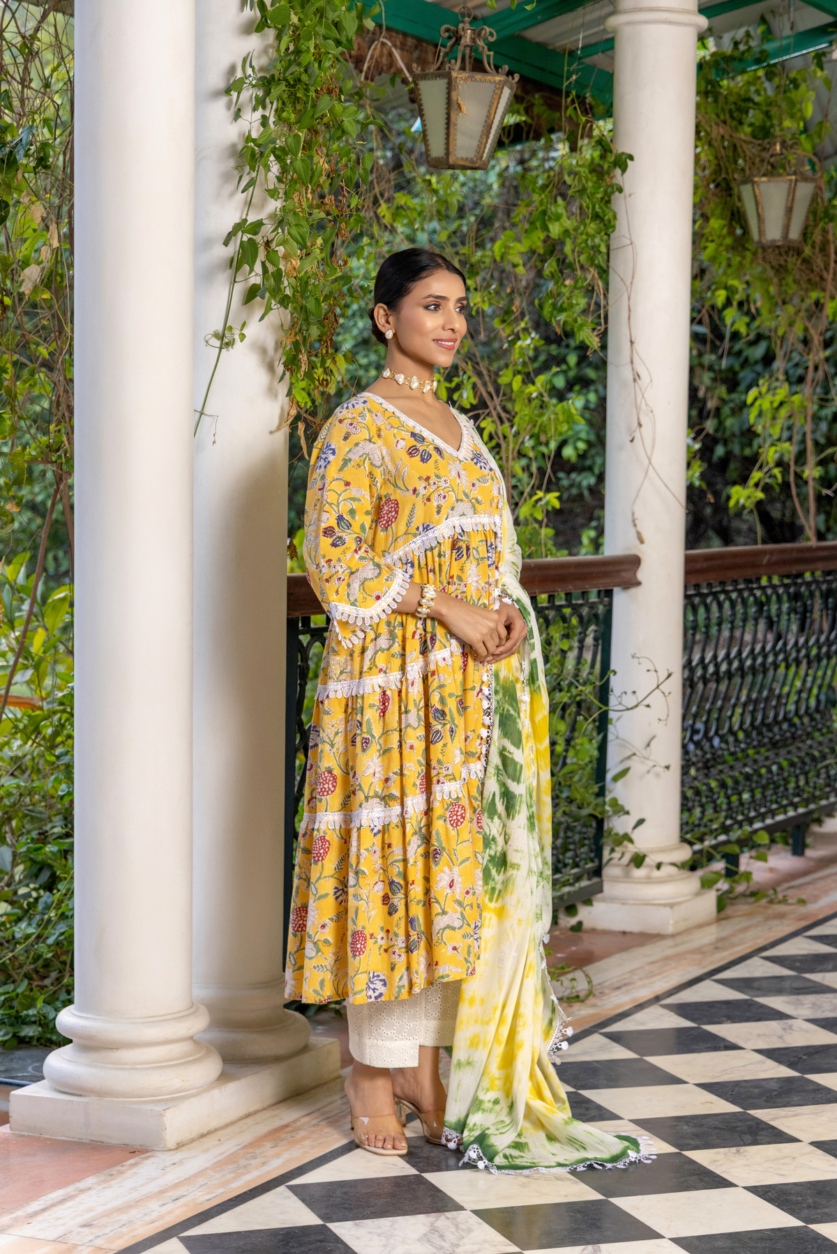 Right side view of a woman stands gracefully in a vibrant yellow floral cotton kurta set, paired with a light green and white dupatta against a lush, green backdrop.