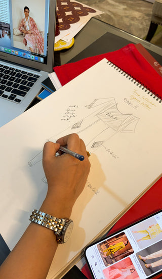A designer sketches a fashion illustration on paper, referencing styles from a laptop and a phone with outfit inspirations.