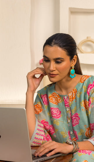 A woman sits at a desk, thoughtfully resting her hand on her chin, wearing a colorful floral kurta and statement earrings, with a laptop in front.