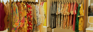 A row of colorful, intricately patterned garments on hangers, featuring bright florals, bold reds, and delicate pastels. The scene feels vibrant and elegant.