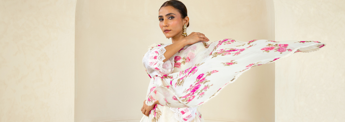 Woman in a white floral outfit poses gracefully against a light beige background, with fabric flowing to the side.