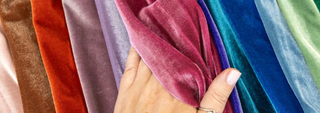 Hand holding and spreading a fan of colorful velvet fabric swatches in shades of red, pink, purple, blue, and green, highlighting the soft texture and sheen.