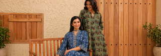 Two women in vibrant, patterned dresses smile in a warm, rustic setting with wooden accents and potted plants, conveying a relaxed and cheerful mood.