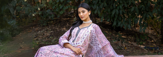 A woman in a floral pink dress sits gracefully outdoors, surrounded by lush greenery. She exudes elegance, wearing a pearl necklace, with a serene expression.