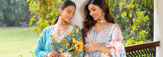 Two women in colorful traditional dresses smiling and standing outdoors near a railing with greenery in the background.