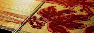 Close-up of red floral embroidery being woven on a loom with threads and needle, with the text “A fabric etched in royal history” visible in the corner.