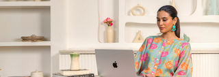 Woman in a colorful floral top works on a laptop in a stylish, minimalist room with white shelves and decorative vases. She appears focused and relaxed.