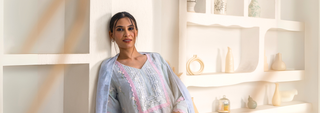 Woman in pastel traditional attire leans against a white textured wall. Background features decorative shelves with vases, creating an elegant and serene tone.
