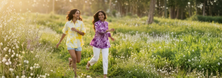 Two women joyfully run through a sunlit meadow filled with wildflowers. One wears a yellow shirt and shorts, the other a purple top and white pants.