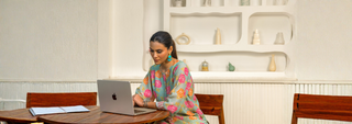 A person works on a laptop at a wooden table, surrounded by an aesthetically pleasing, minimalist white interior with decorative items.