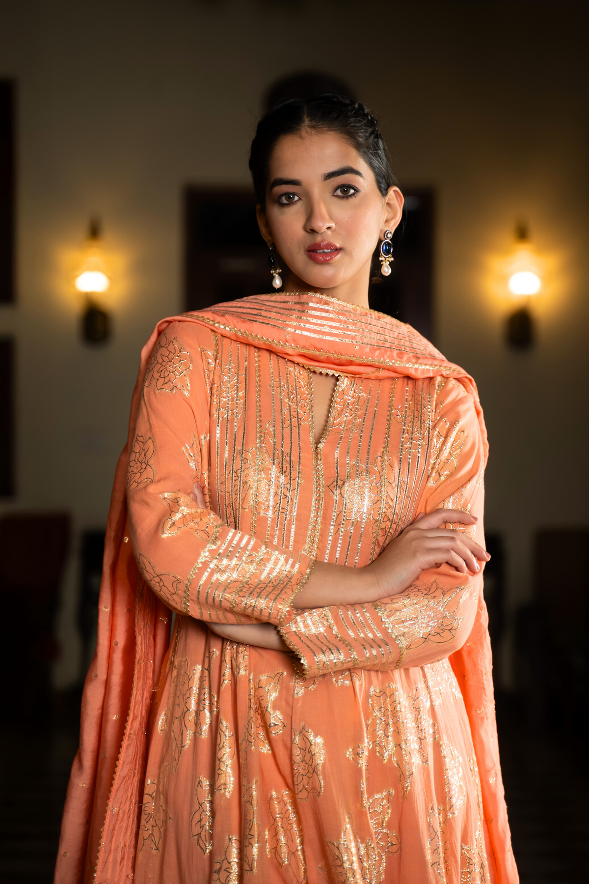 Closeup view of a  woman stands elegantly in an orange, traditional anarkali kurta set showcasing intricate embroidery against a beautifully decorated interior.