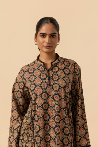A woman wearing a long-sleeve ajrakh co-ord set with an intricate geometric and floral pattern in brown, rust, and black, set against a solid beige background.