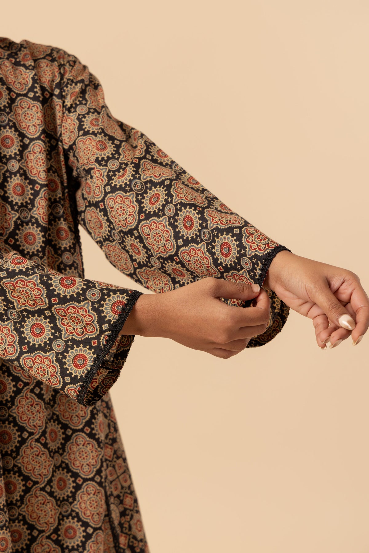 A close-up of a person wearing a long-sleeve patterned ajrakh co-ord set with a black base and intricate orange, beige, and red floral motifs, shown against a solid beige background.
