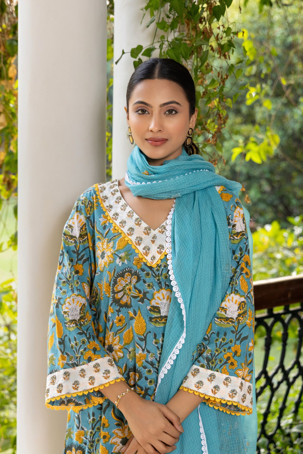 A person wears a vibrant turquoise and yellow handblock printed kurta set with a dupatta rounder on her neck, standing gracefully on a checkered floor amidst lush greenery.