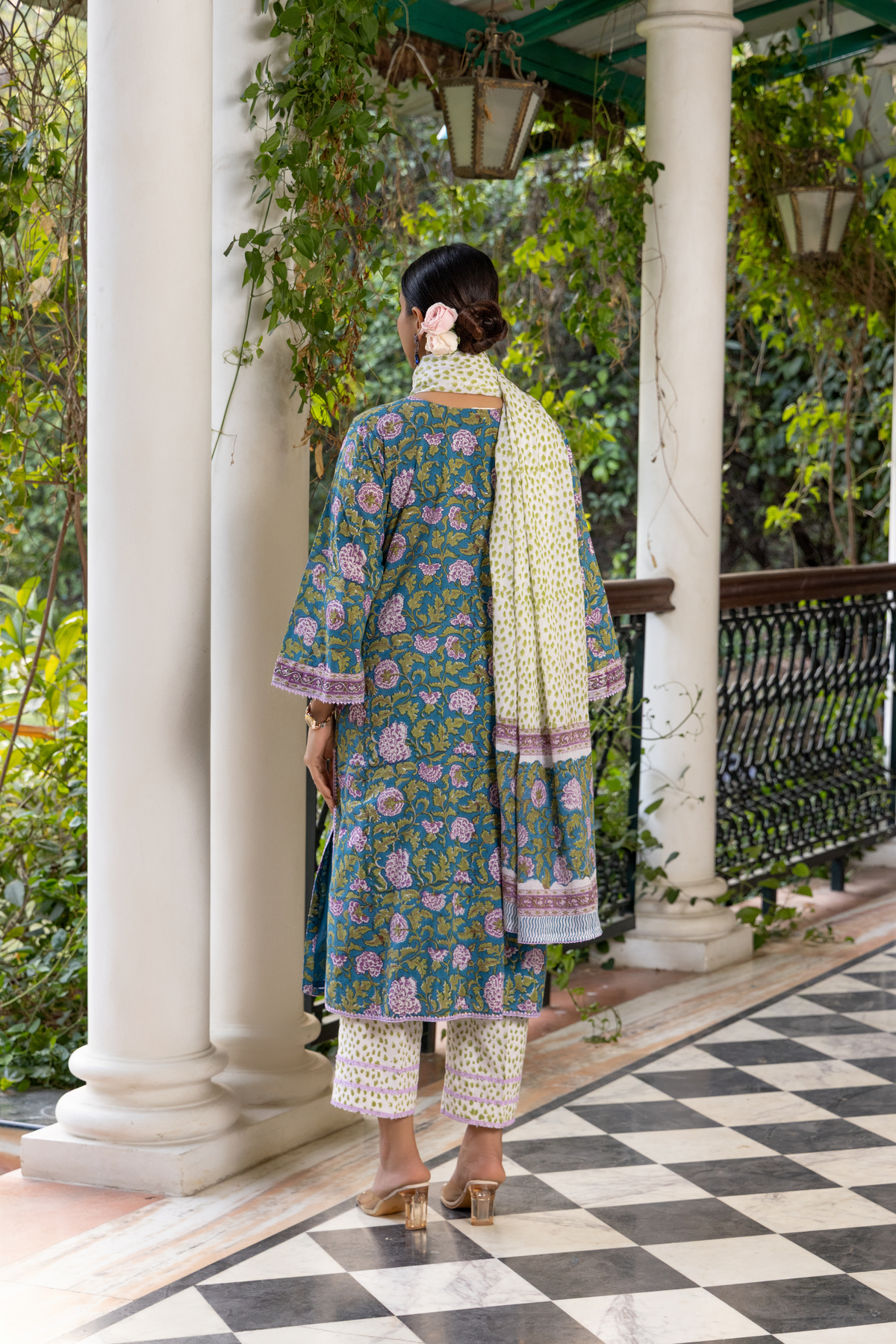 Back view of  a woman wears a colorful, peacock-printed cotton kurta set with a matching scarf, standing on a checkered floor surrounded by greenery.