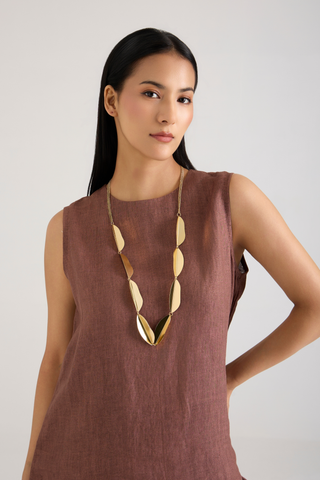 A woman wearing a sleeveless dusty rose linen top and a chunky gold geometric pendant necklace, photographed against a neutral light gray background.