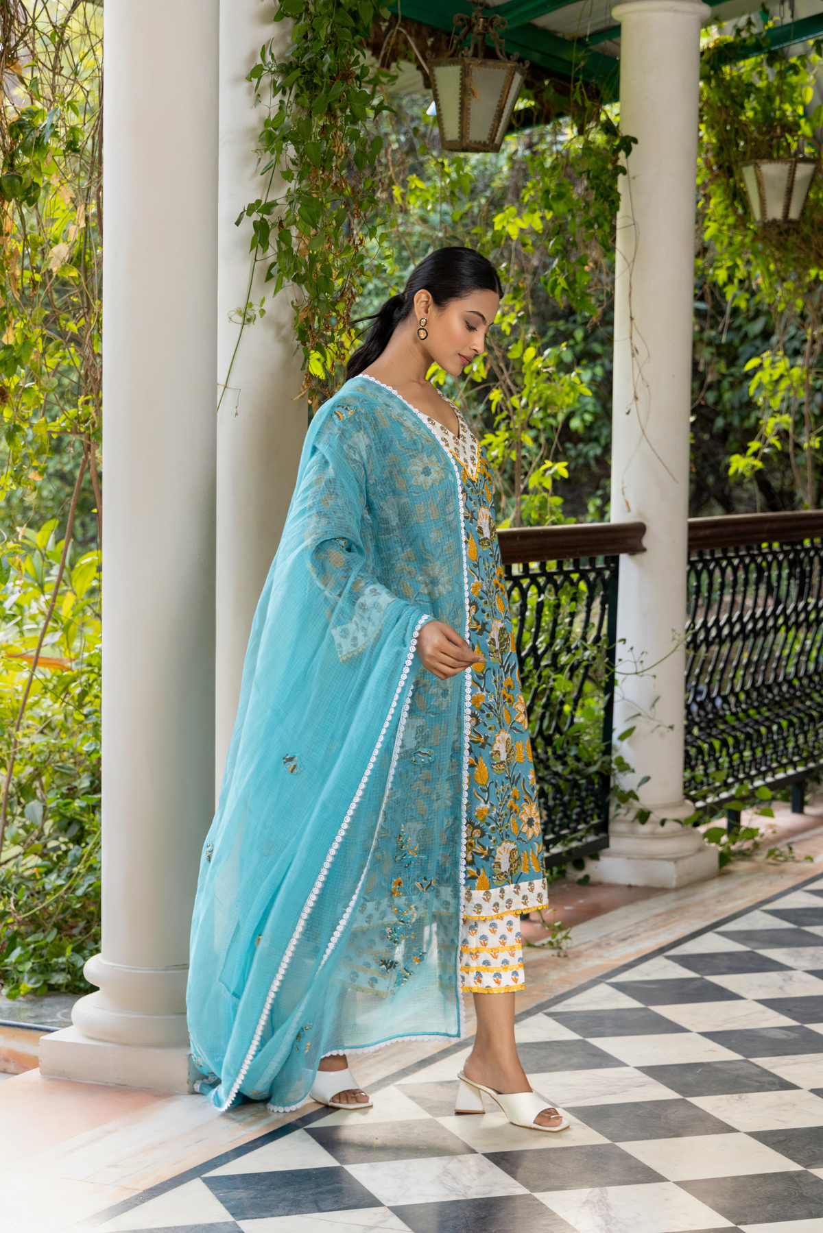 Right side view of a  person wears a vibrant turquoise and yellow handblock printed kurta set, standing gracefully on a checkered floor amidst lush greenery.