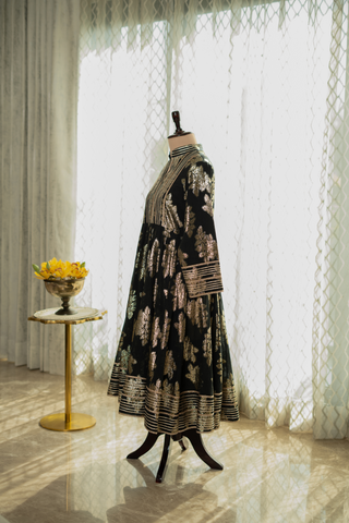 Side view of black Anarkali suit with intricate gold embroidery, displayed on a mannequin against a softly lit curtain backdrop.