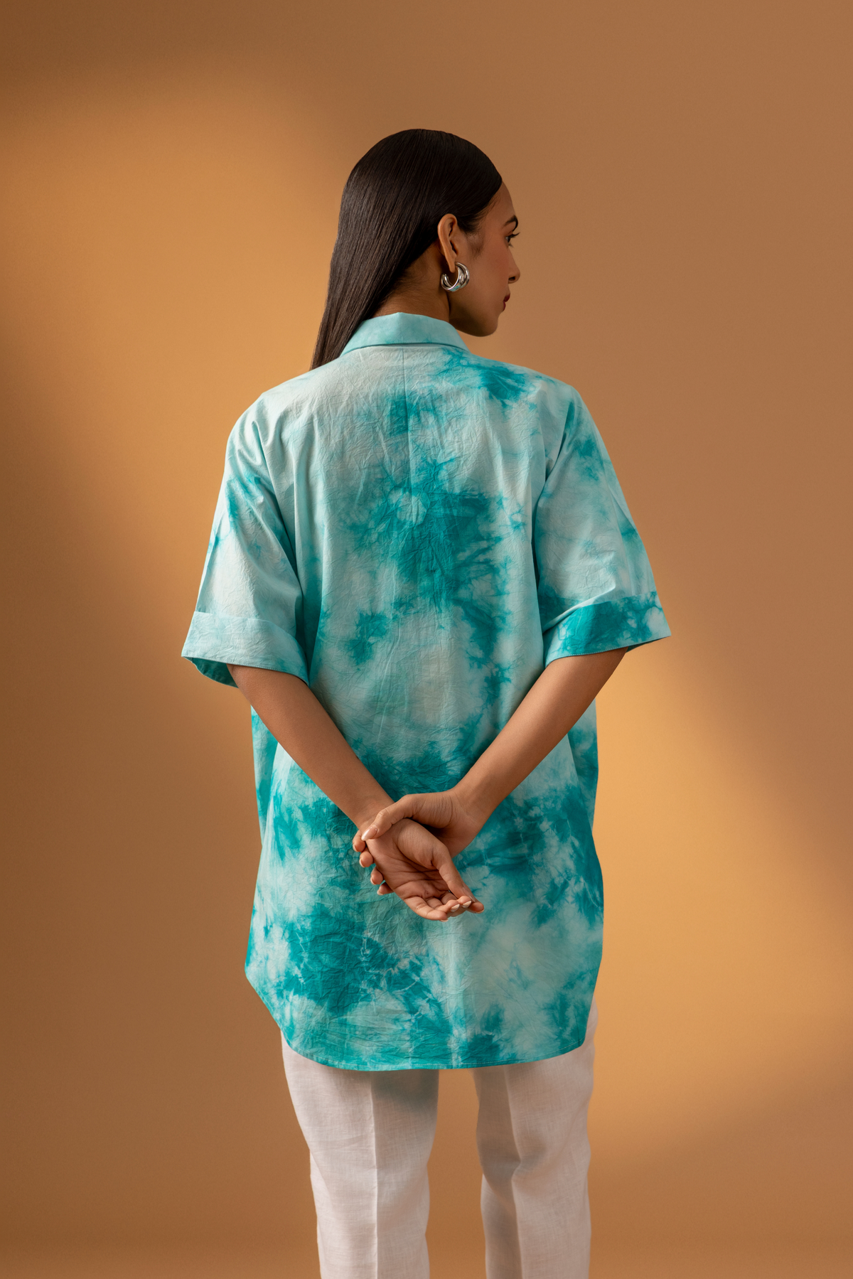 Back view if a woman stands confidently in a teal tie-dye oversized shirt and light-colored trousers against a warm, neutral background.