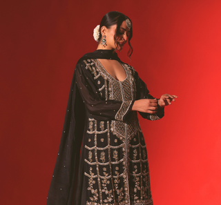 A woman modeling a black traditional embroidered dress with intricate silver mirror work and a matching sheer black dupatta, posed against a solid red studio background.
