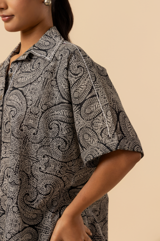 A model stands confidently in a matching patterned outfit, featuring a loose shirt stands against a soft beige background, showcasing a relaxed style.