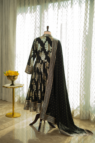 back view of black Anarkali suit with intricate gold embroidery, paired with a matching dupatta featuring delicate gold accents, displayed on a mannequin against a softly lit curtain backdrop.