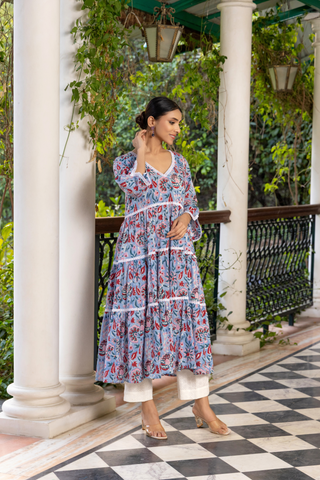 Right side tilt view of a woman stands gracefully on a checkered marble floor, wearing a floral patterned handblock printed kurta set dress, surrounded by lush greenery and elegant columns.