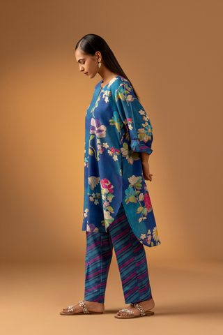 Side view of a woman models a vibrant floral co-ord set paired with striped pants, set against a warm, neutral background.
