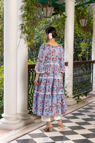 Back view of a woman stands gracefully on a checkered marble floor, wearing a floral patterned handblock printed kurta set dress, surrounded by lush greenery and elegant columns.