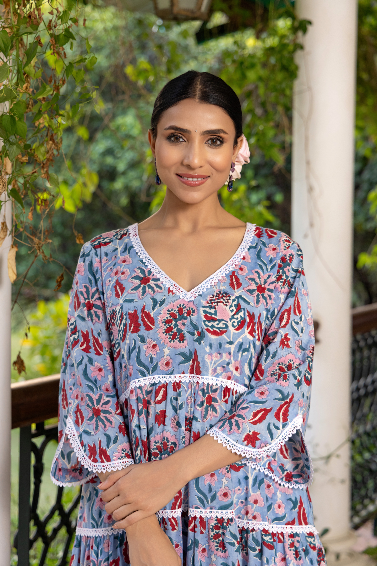 Closeup view of a woman stands gracefully on a checkered marble floor, wearing a floral patterned handblock printed kurta set dress, surrounded by lush greenery and elegant columns.